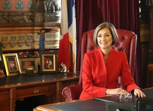 Image of Governor Kim Reynolds seated behind the official desk, smiling and wearing a red suit